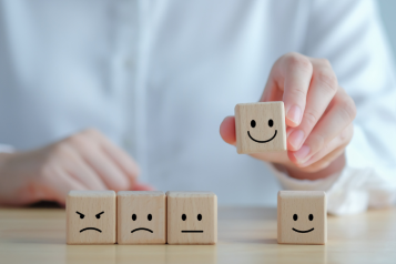 A hand lifting a wooden blocks with a smiley face and the table has other blocks with other emotions